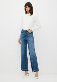 Spodnie dżinsowe o szerokich nogawkach w kolorze niebieskim, ozdobione perłami, w połączeniu z dopasowaną, białą bluzką z długim rękawem. Stylizacja charakteryzuje się nowoczesnym, minimalistycznym designem.