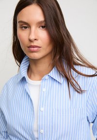 Plava i bijela prugasta košulja na kopčanje s špicastim ovratnikom, prednjim džepom i mekanom teksturom od pamuka, nošena preko bijele majice.