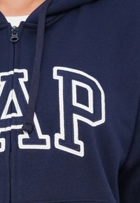 Navy zip-up hoodie i blandning av bomullsfabric; har vita broderade "GAP"-bokstäver på bröstet och snörning i huvan.