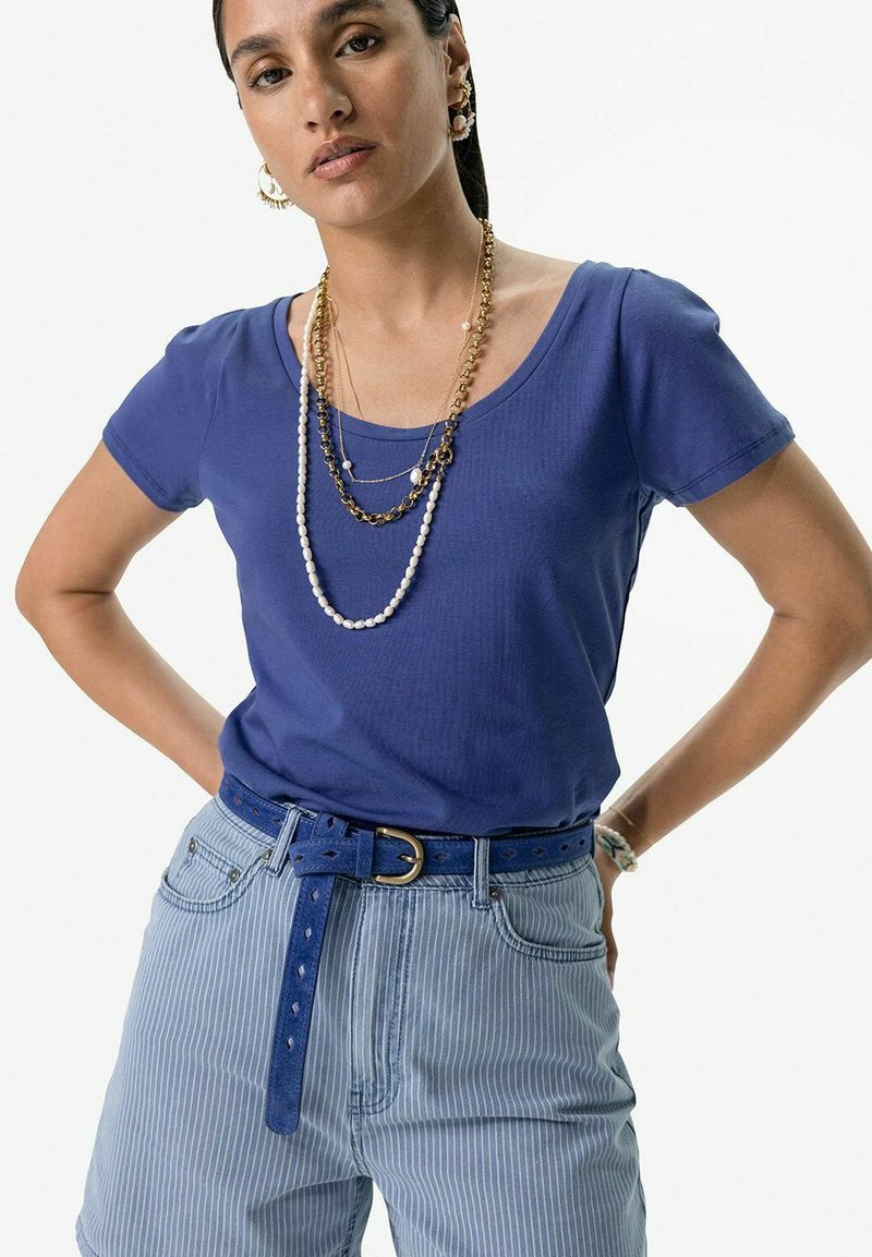 T-shirt bleu à manches courtes avec un col rond, associé à un short rayé bleu clair et une ceinture bleue. Colliers superposés en or et perles.