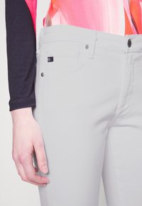 Valkoiset puuvillafarkut, joissa on kapea malli, etutasku, metalliset napin yksityiskohdat ja pieni brändilappu vyötäröllä.