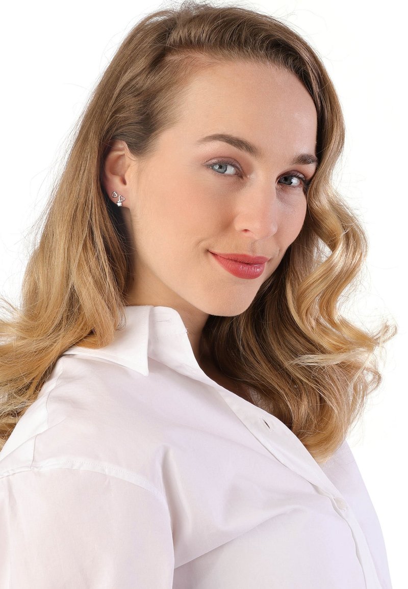 Zilveren hartvormige studie oorbellen gedragen door een persoon met lichtbruin golvend haar, gekleed in een witte blouse met kraag.