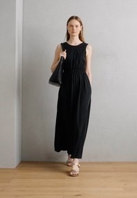 Abito nero senza maniche con vita elastica, design lungo fino al pavimento, abbinato a una borsa nera e sandali leggeri su un pavimento in legno.