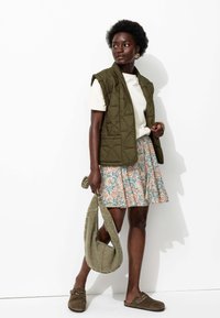 Gilet matelassé vert olive sur un t-shirt blanc, associé à une jupe à fleurs avec des motifs dans des tons bleus et pêches. Les accessoires comprennent un sac vert.