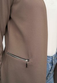 Cardigan marron texturé avec une poche zippée, présentant une fermeture éclair lisse et un design structuré. Porté sur une chemise claire et un jean en denim.