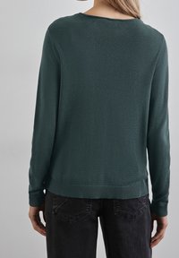 Mörkgrön tröja med långa ärmar, rund halsringning, ribbmuddar i nederkant och ärmar, tillverkad av ett mjukt tyg, bärs över mörka jeans.