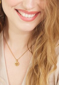 Gouden ketting met ronde hanger met een ontwerp van de letter "K". De ketting is delicaat en de schakels zijn zichtbaar tegen een lichte huid.