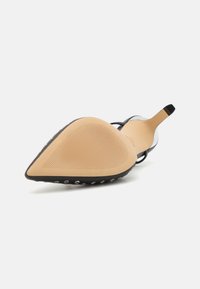 Escarpin noir à bout pointu avec une bride transparente, semelle beige dotée de stries et accents argentés sur les bords pour une meilleure adhérence.