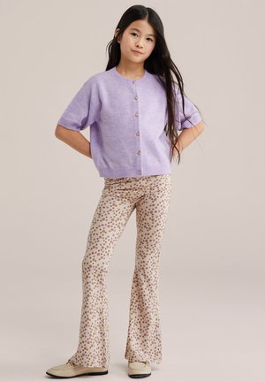 Pikkade tumejate juustega tüdruk kannab lavendlivärvi nööbitud kardigani ja beeži lillemustriga laiade säärtega pükse, seisab käed puusadel.