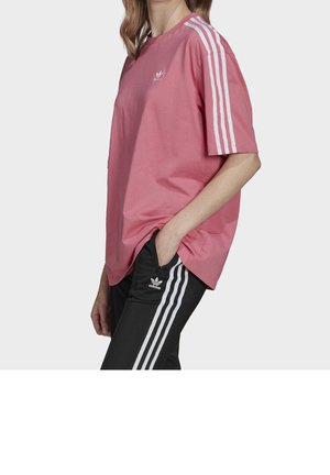 Personne portant un T-shirt Adidas rose avec des bandes blanches sur les manches et un pantalon Adidas noir avec des bandes blanches, la main dans la poche.