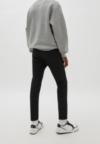 Personne debout portant un sweat-shirt gris, un pantalon noir ajusté, des chaussettes blanches et des baskets blanches avec des accents noirs sur un fond blanc uni.