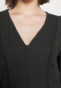 Svart blus med V-ringning, texturerat tyg, dekorativa knappar fram och volangärmar, med spetsdetaljer på sidorna.