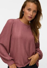 Blouse rose en tissu texturé, avec un col rond et des manches longues bouffantes. Présente un motif crêpé subtil et une coupe décontractée.