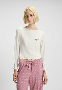 Witte lange mouwen top met een golvende zoom en "HUGO" logo. In combinatie met roze pyjamabroek met een herhaald logo-patroon en trekkoord.