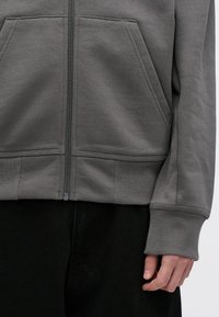 Siva zip-up kapuca z teksturirano tkanino, sprednjima žepoma in rebrastimi manšetami, v kombinaciji s črnimi hlačami. Podroben pogled na detajle oblačila.