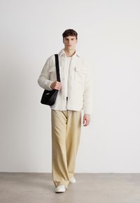 Hvit fleece skjortejakke over en hvit topp, parret med beige vide bukser og hvite sneakers, bærende en svart crossover-veske.