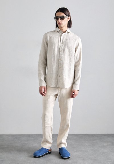 Chemise en lin beige clair avec fermeture à boutons, pantalon en lin assorti et sabots bleus. Les lunettes sont carrées avec des montures épaisses.