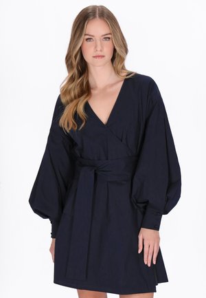 Giovane donna con capelli biondi mossi che indossa un abito avvolgente blu navy con maniche lunghe a sbuffo e cintura annodata in vita, in piedi su uno sfondo bianco.