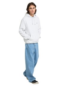 Jeune homme aux cheveux bruns portant un sweat à capuche blanc et un jean bleu large, debout les mains dans la poche du sweat sur fond blanc.