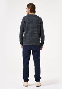 Dunkel strukturierter Strickpullover mit rundem Ausschnitt, kombiniert mit marineblauen Hosen. Die Schuhe sind hellfarbige, lässige Sneakers.