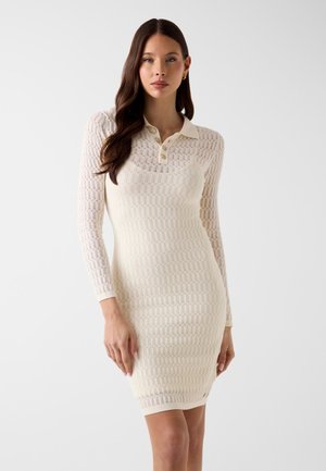 Femme portant une robe en maille crème, ajustée, à manches longues, avec un col et des boutons, debout devant un fond blanc uni.