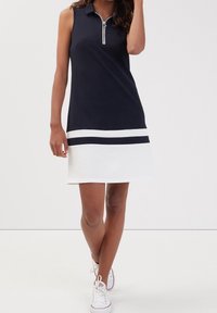 Robe polo sans manches bleu marine avec une fermeture éclair, une bande blanche en bas, et une bande horizontale contrastante. Portée avec des baskets blanches. Texture de tissu lisse.