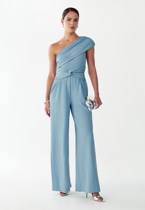 Žena v svetlomodrom jednoramennom overal so širokými nohavicami, strieborné vysoké podpätky, strieborné listové kabelka a minimalistické šperky.