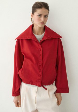 Junge Frau trägt eine rote Jacke mit breitem Kragen und Knöpfen über einem hellgrauen Hemd und cremefarbenen Hosen, stehend vor einem einfarbigen Hintergrund.