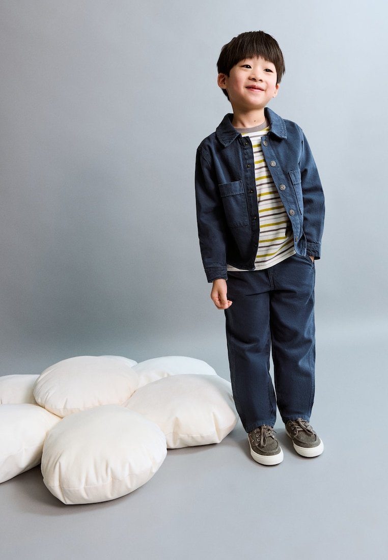 Jeune garçon portant une chemise rayée, une veste et un pantalon bleu marine, debout près de gros coussins blancs ronds sur un sol et un mur gris.