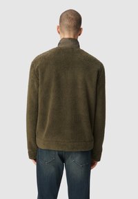 Veste verte olive duveteuse avec une texture douce, présentant un design simple et un col rond, associée à un jean en denim bleu.