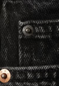 Detailní pohled na černou denimovou látku, která obsahuje černý patentní knoflík a měděný knoflík s vyrytým nápisem "WEEKDAY", s texturovaným povrchem.