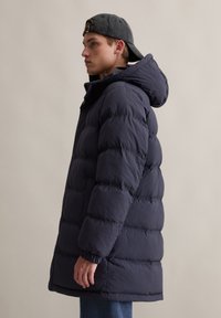Dunkelblaue Daunenjacke mit Kapuze, die ein gestepptes Design, elastische Bündchen und einen geraden Schnitt aufweist. Über blauen Jeans getragen.