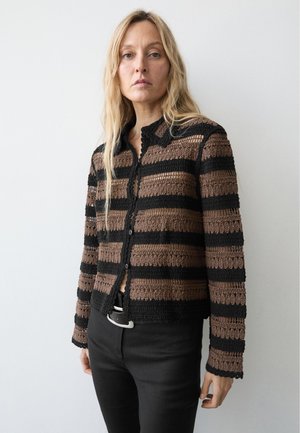 Vrouw met lang blond haar die een bruin-zwart gestreepte, gestructureerde gebreide cardigan en zwarte broek draagt tegen een effen witte achtergrond.