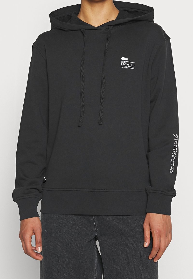 Homme portant un sweat à capuche Lacoste noir avec un logo texte blanc sur la poitrine et la manche, associé à un jean foncé, debout devant un fond uni.