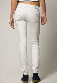Jean skinny en denim blanc avec poches arrière, présentant un design épuré et une texture subtile. Associé avec des bottes à chevilles en daim bleu.