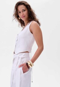 Biała, krótkotaliowa kamizelka z dekoltem w szpic, zdobiona guzikami, zestawiona z białymi spodniami o wysokim stanie, związanymi w talii. Złote i brązowe bransoletki zdobią nadgarstek.