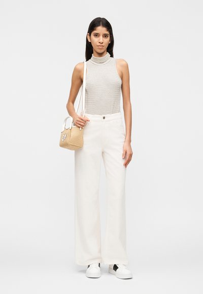 Γυναίκα με αμάνικη ριγέ ζιβάγκο και φαρδύ λευκό παντελόνι, κρατώντας μικρή ψάθινη τσάντα, στέκεται μπροστά σε απλό φωτεινό φόντο.