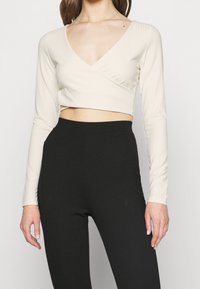 Hellbeiges, geripptes, langärmliges, kurz geschnittenes Oberteil mit V-Ausschnitt und Wickeldesign, kombiniert mit hochtaillierten schwarzen Leggings.