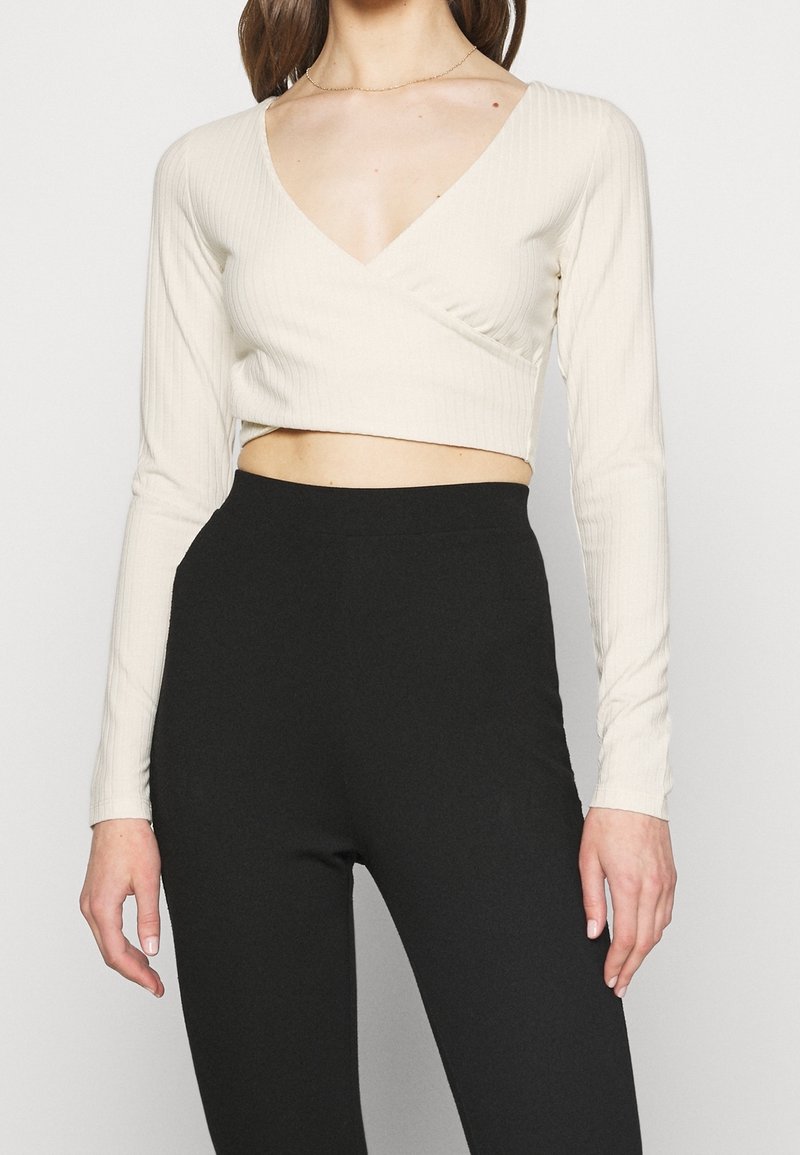 Hellbeiges, geripptes, langärmliges, kurz geschnittenes Oberteil mit V-Ausschnitt und Wickeldesign, kombiniert mit hochtaillierten schwarzen Leggings.