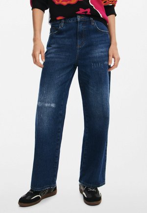 Jeans a gamba larga in denim blu con vita alta, caratterizzati da leggere scollature e tasche standard. Indossati con scarpe nere a fantasia.