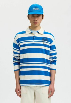 Jeune homme portant un polo à manches longues rayé bleu et blanc et une casquette bleue assortie, sur fond blanc uni.