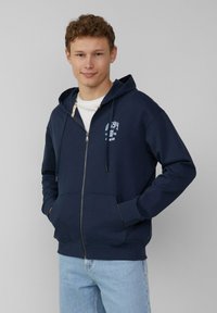 Felpa con zip di colore blu navy realizzata in tessuto morbido, con tasca a marsupio, cappuccio regolabile con stringhe e dettaglio del logo stampato bianco sul petto.