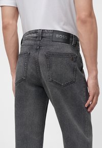Homme portant un jean en denim gris foncé avec des poches arrière et une étiquette en cuir noire indiquant "BOSS" sur la ceinture.