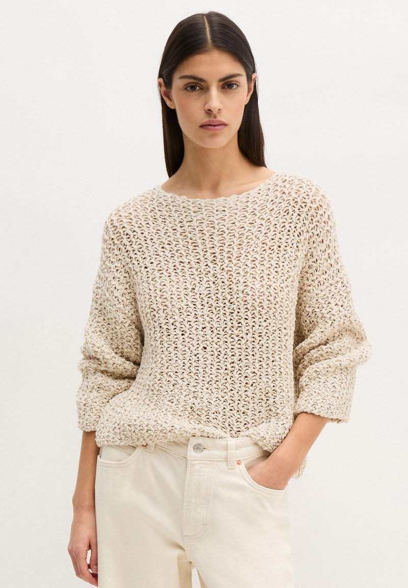 Femme aux longs cheveux noirs portant un pull beige en maille large et un pantalon de couleur claire, debout devant un fond uni.