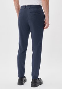 Homme portant un pantalon bleu marine slim avec des poches arrière boutonnées et des chaussures noires, debout devant un fond clair uni.