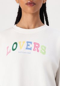 Fehér pulóver, melyen színes hímzett betűk formálják a "LOVERS" szót, alatta pedig a "Self Care Club" felirat található. Puha, könnyű anyag, kerek nyakkivágással.