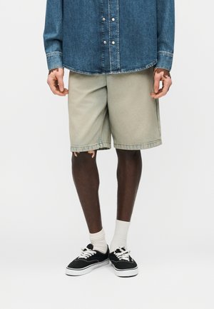 Personne portant un short beige clair, une chemise en jean bleue boutonnée, des chaussettes blanches et des baskets noires sur fond blanc.