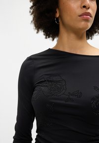 Sieviete, ģērbusies melnā garās piedurkņu kreklā ar melniem rozā rakstiem un sudraba riņķiem ausīs, redzama no kakla līdz jostasvietai.