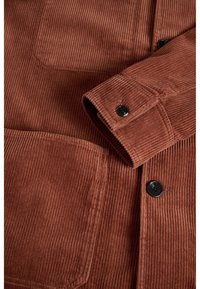 Close-up van een roestkleurige corduroy jas mouw, met zwarte knopen en een groot voorvak.