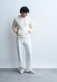 Moški z kodrastimi lasmi, oblečen v svetlo bež kapucar, široke bele hlače in bele superge, stoji ob bledo sivem ozadju z rokami v žepih.